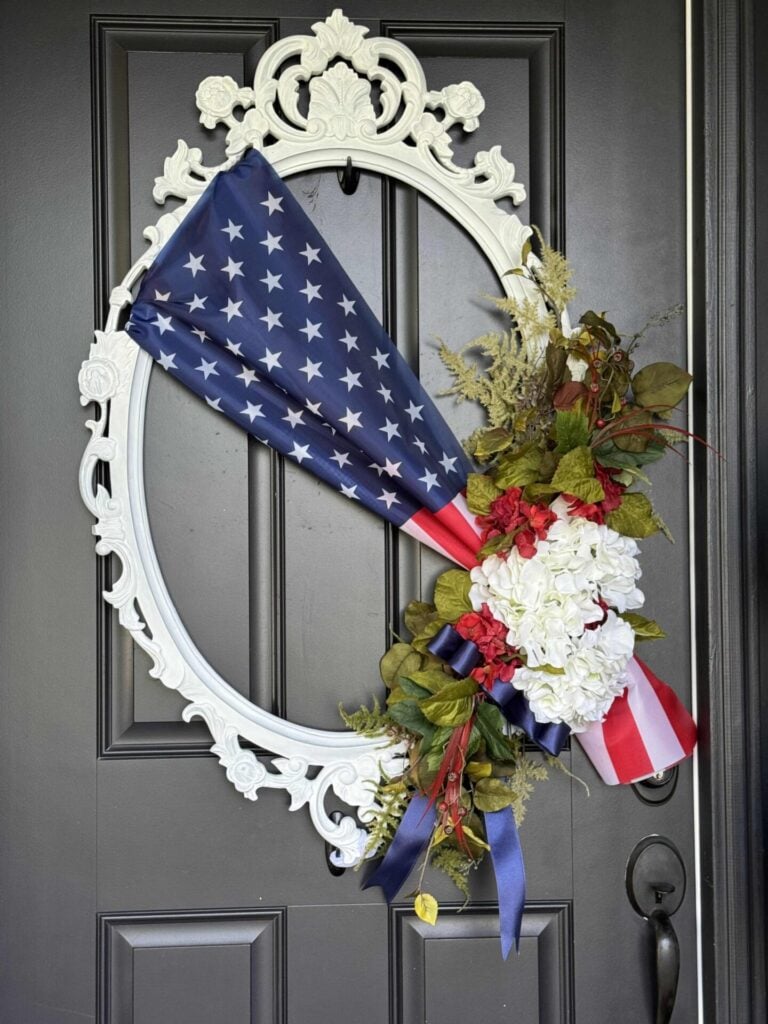 A black door displays a white ornate oval frame wreath decorated with an American flag, white hydrangeas, red flowers, greenery, and blue ribbon—perfect 4th of July front door decor for a patriotic and floral arrangement.