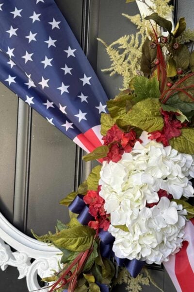 A patriotic wreath featuring white hydrangeas, red flowers, green leaves, and an American flag ribbon makes the perfect 4th of July front door decor on a black door with a decorative white frame.