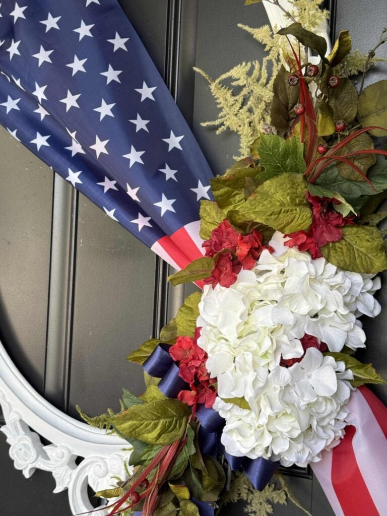 A patriotic wreath featuring white hydrangeas, red flowers, green leaves, and an American flag ribbon makes the perfect 4th of July front door decor on a black door with a decorative white frame.