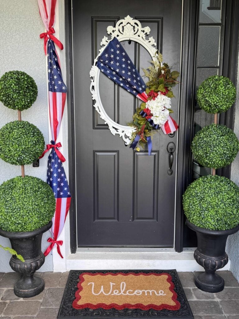 A black front door decorated with a patriotic wreath featuring American flag fabric and red, white, and blue ribbons. Two topiary plants sit on each side, and a welcome mat lies at the doorstep.