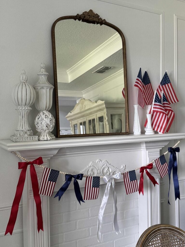 A fireplace mantel decorated with a DIY patriotic garland made with small American flags, red, white, and blue ribbons, and white decorative vases. A large mirror reflects part of the room’s elegant decor.
