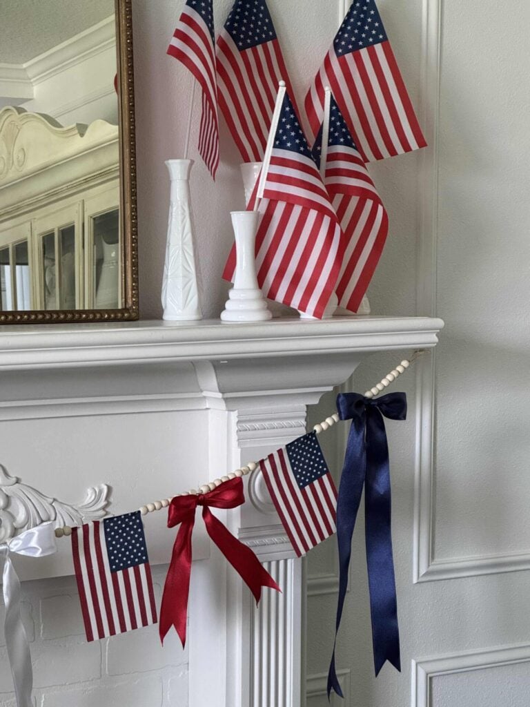 A white mantel decorated with small American flags in white vases and a garland of mini flags and red and blue ribbon bows. A mirror is partially visible above the mantel.