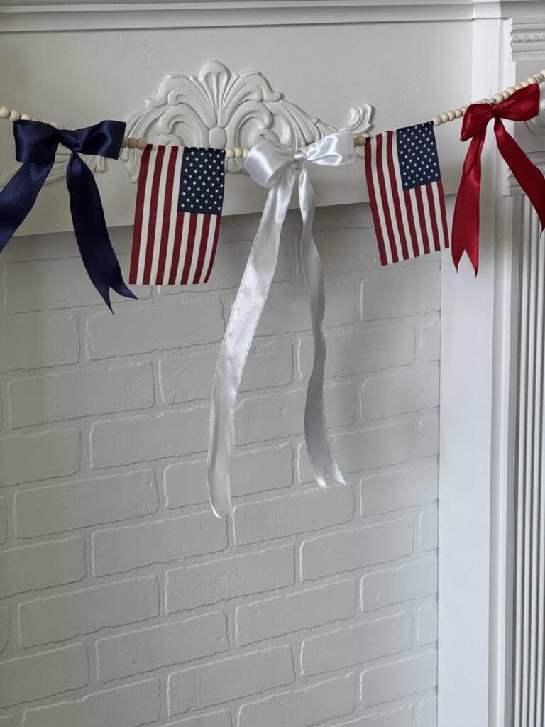 A white mantel decorated with a garland of small American flags and red, white, and blue ribbon bows, hanging against a white brick wall.