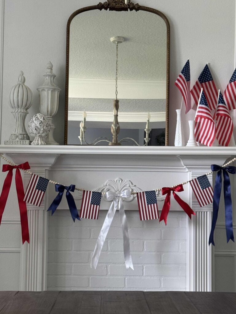 A fireplace mantel decorated with small American flags in vases, DIY Patriotic Ribbon Flags, red, white, and blue ribbons, and a garland of mini flags. A large mirror and decorative white objects sit on the mantel.