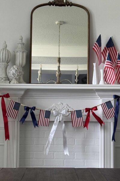 A white fireplace mantel decorated with small American flags, a garland of red, white, and blue bows and flags, and decorative vases. A large mirror hangs above, reflecting a chandelier.