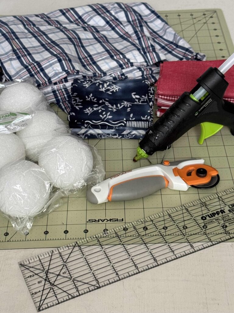 Craft supplies arranged on a cutting mat, including fabric squares, white foam balls, a rotary cutter, a clear ruler, and a black hot glue gun. The items are ready to make DIY fabric orbs. 