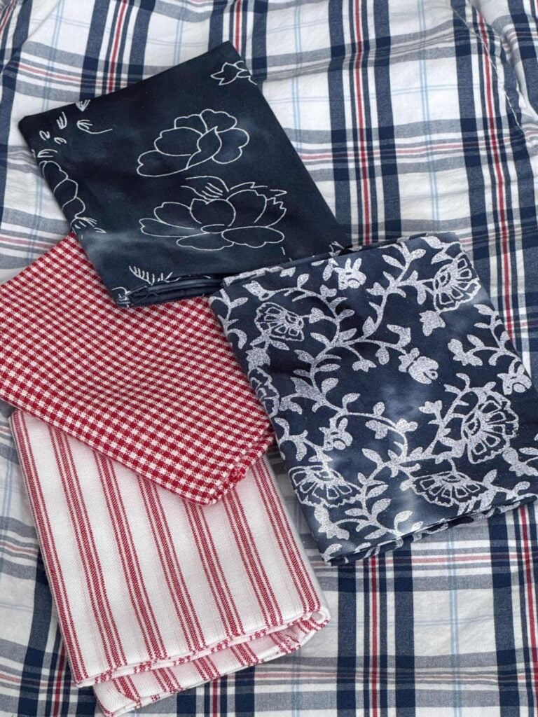 Four folded fabrics lay on a blue and white plaid cloth: two are red (one with stripes, one with checks), and two are dark blue with white floral patterns. To be used to make DIY fabric orbs. 
