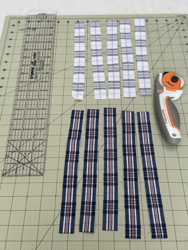 A cutting mat with a clear quilting ruler, a rotary cutter, and eight plaid fabric strips—four in white with blue and red lines, and four in navy with white and red lines—arranged in parallel rows. Strips will be used to make DIY fabric orbs. 