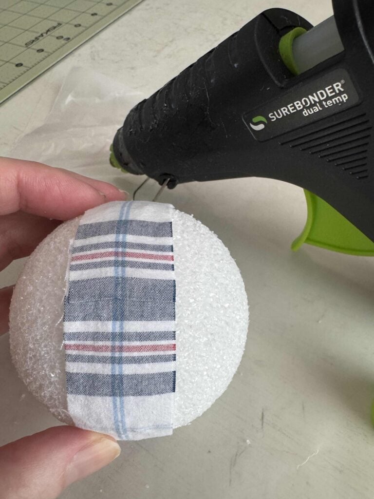 A hand holds a white foam ball with a strip of plaid fabric glued on it; a black hot glue gun and crafting supplies are visible in the background.