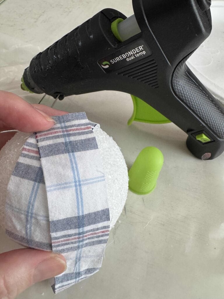 A hand holds a piece of plaid fabric against a white foam ball, with a hot glue gun and a green finger protector visible on a white surface in the background.
