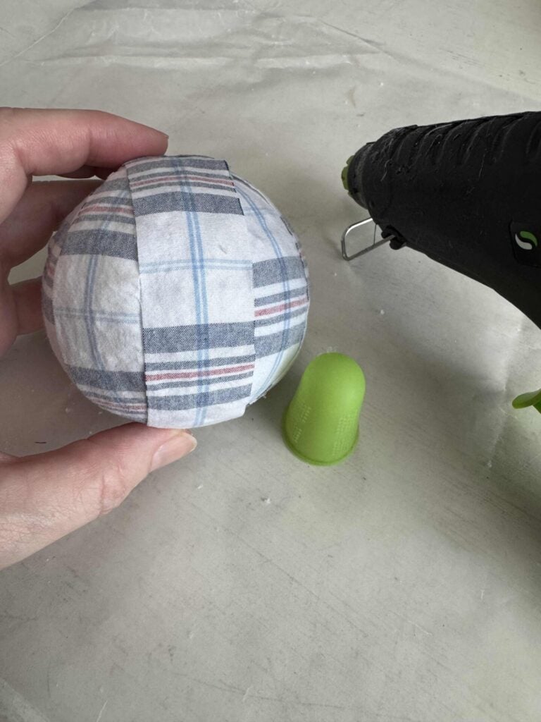 A hand holds a foam ball wrapped in plaid fabric strips. Nearby are a green thimble and a black hot glue gun on a white surface.