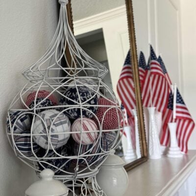 A decorative white wire basket filled with red, white, and blue DIY Fabric Orbs sits on a mantel, with white vases and several small American flags in the background, reflecting a patriotic theme.