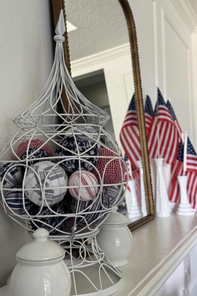 A decorative white wire basket filled with red, white, and blue DIY Fabric Orbs sits on a mantel, with white vases and several small American flags in the background, reflecting a patriotic theme.