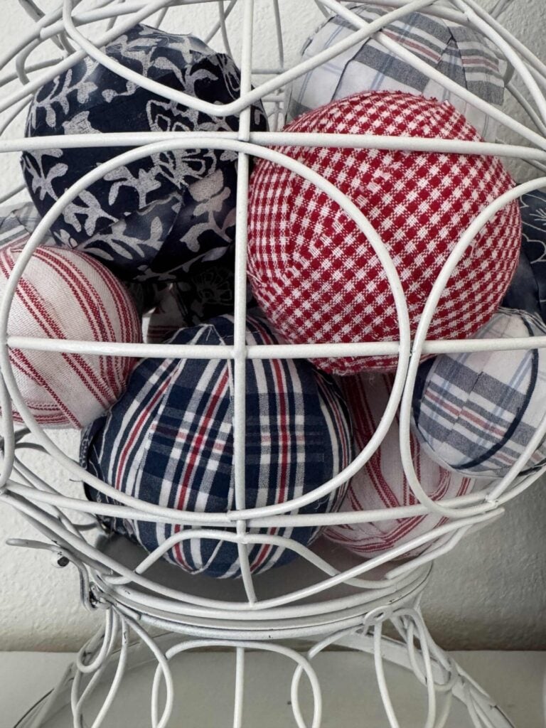 A white wire basket holds several DIY Fabric Orbs—fabric-covered balls in red, white, and blue plaid, gingham, and floral patterns—arranged closely together.