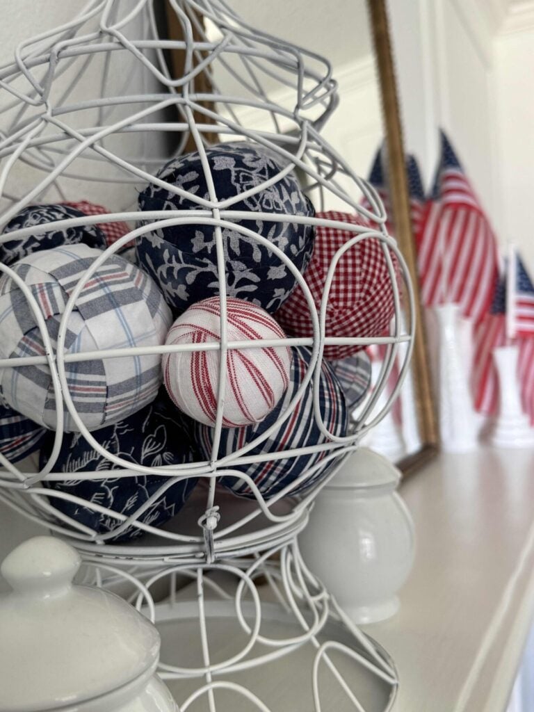 A white wire basket holds fabric balls with red, white, and blue patterns. In the background, several small American flags and a DIY Patriotic Ribbon Flag are displayed on a mantel with white decor items.