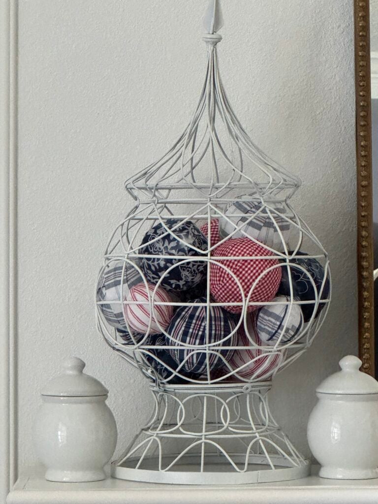 A decorative white wire cage filled with red, white, and blue patterned fabric balls sits on a shelf between two white ceramic containers with lids, perfect as an accent for a DIY patriotic garland display.