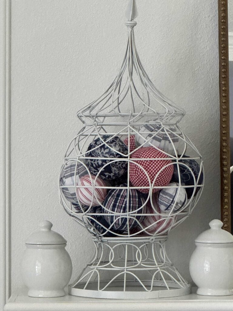 A decorative white wire cage filled with DIY Fabric Orbs in red, white, and blue patterns sits between two white ceramic jars on a white shelf against a light-colored wall.