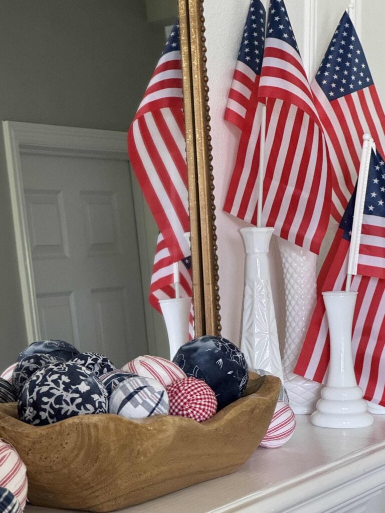 A wooden bowl of red, white, and blue DIY Fabric Orbs sits on a mantel beside white vases holding American flags, with a mirror reflecting the flags and part of a white door in the background.
