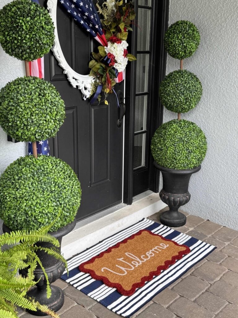 A black front door with a patriotic wreath, 4th of July front door decor accents, two topiary plants in black urns, a striped “Welcome” doormat, and potted greenery on a stone porch.