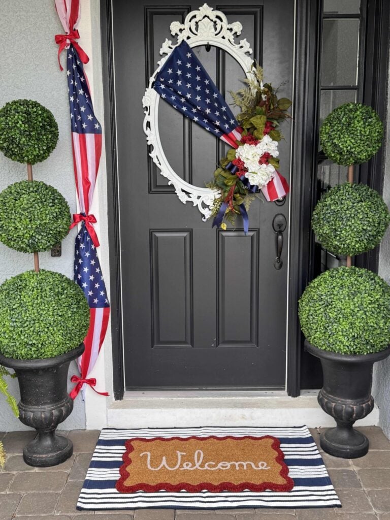 Black front door with a white oval wreath decorated with an American flag and flowers—perfect 4th of July front door decor. Two potted topiary plants flank the door, and a striped “Welcome” mat sits at the doorstep. Another flag is draped to the left.
