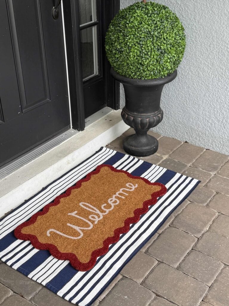 A brown doormat with “Welcome” in white script is placed on a striped rug in front of a black door, creating a perfect base for your 4th of July front door decor. Nearby, a round topiary plant sits in a black planter on the stone patio.