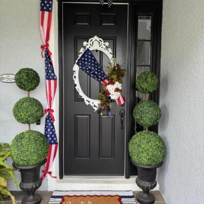 A black front door with 4th of July front door decor, featuring a patriotic wreath and American flag swag. Two potted topiary plants flank the “Welcome” mat atop a blue and white striped rug. The house exterior is light gray.