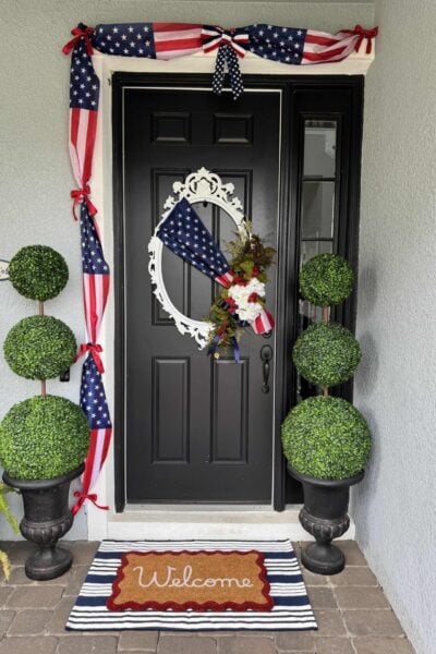 A black front door with 4th of July front door decor, featuring a patriotic wreath and American flag swag. Two potted topiary plants flank the “Welcome” mat atop a blue and white striped rug. The house exterior is light gray.