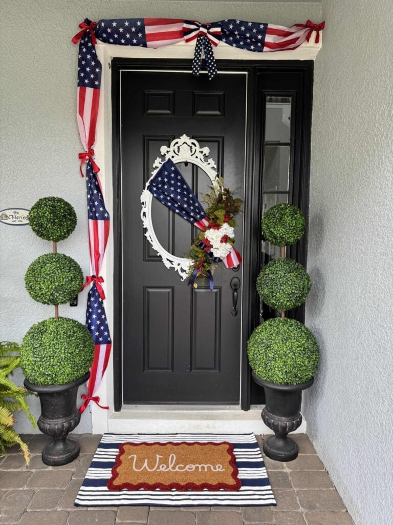 A black front door with 4th of July front door decor, featuring a patriotic wreath and American flag swag. Two potted topiary plants flank the “Welcome” mat atop a blue and white striped rug. The house exterior is light gray.