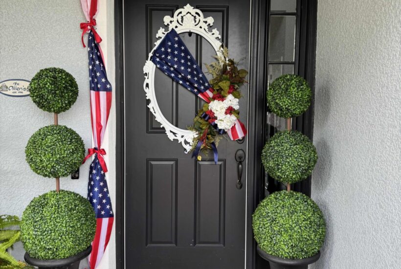 A black front door with 4th of July front door decor, featuring a patriotic wreath and American flag swag. Two potted topiary plants flank the “Welcome” mat atop a blue and white striped rug. The house exterior is light gray.