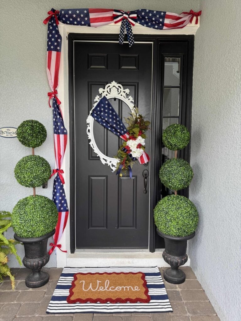 A black front door with a white wreath and patriotic bow, decorated with American flag ribbons and a small bouquet of flowers. Two topiary plants flank the entrance, and a Welcome mat sits on a striped rug.