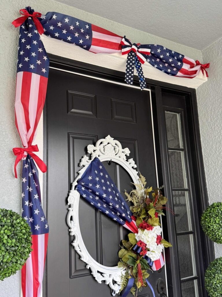 A black front door is adorned with 4th of July front door decor, featuring American flag-themed fabric, red bows, and a white ornate wreath holding a bouquet of flowers. Two green topiary plants sit proudly on either side.