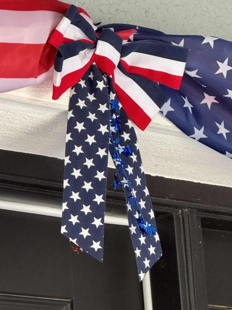 A decorative bow made from American flag-themed ribbon, perfect as 4th of July front door decor, features red, white, and blue stripes with white stars on a blue background and is tied above a doorway.