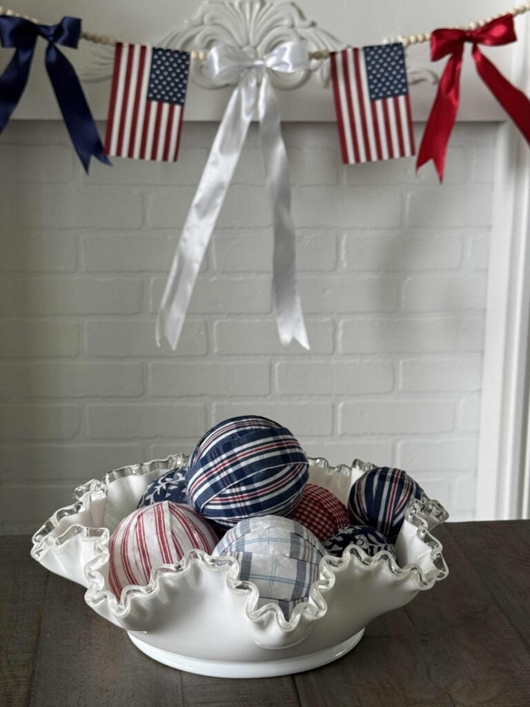 A white ruffled bowl filled with DIY fabric orbs in red, white, and blue patterns sits on a table. Behind it, small American flags and red, white, and blue ribbons are hung as festive decor.