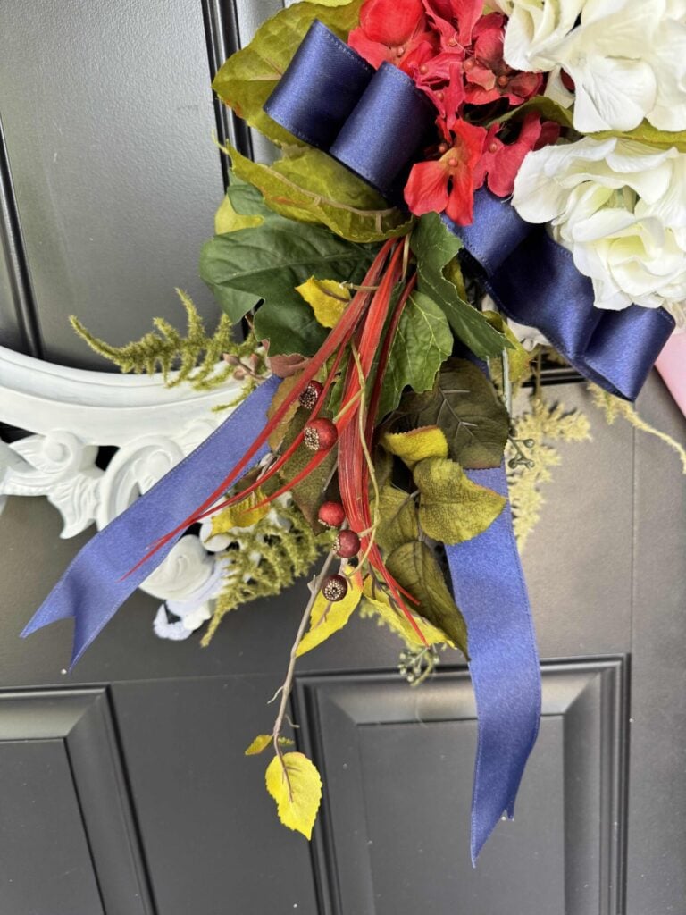 A close-up of a door wreath with deep blue ribbon, red and white flowers, green leaves, yellow foliage, and small acorns on a dark door. Part of a white decorative frame is visible in the background.