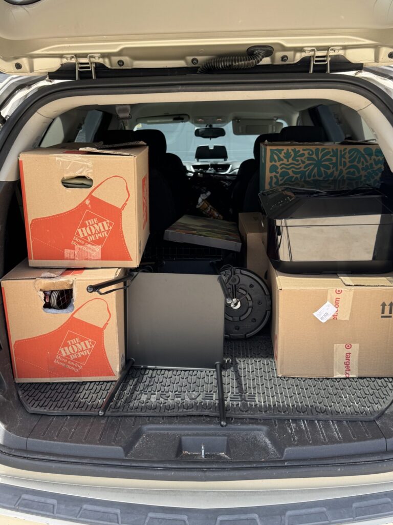 The open trunk of an SUV loaded with cardboard moving boxes, a small teal-patterned bin, a black printer, and various household items, indicating a move or relocation.