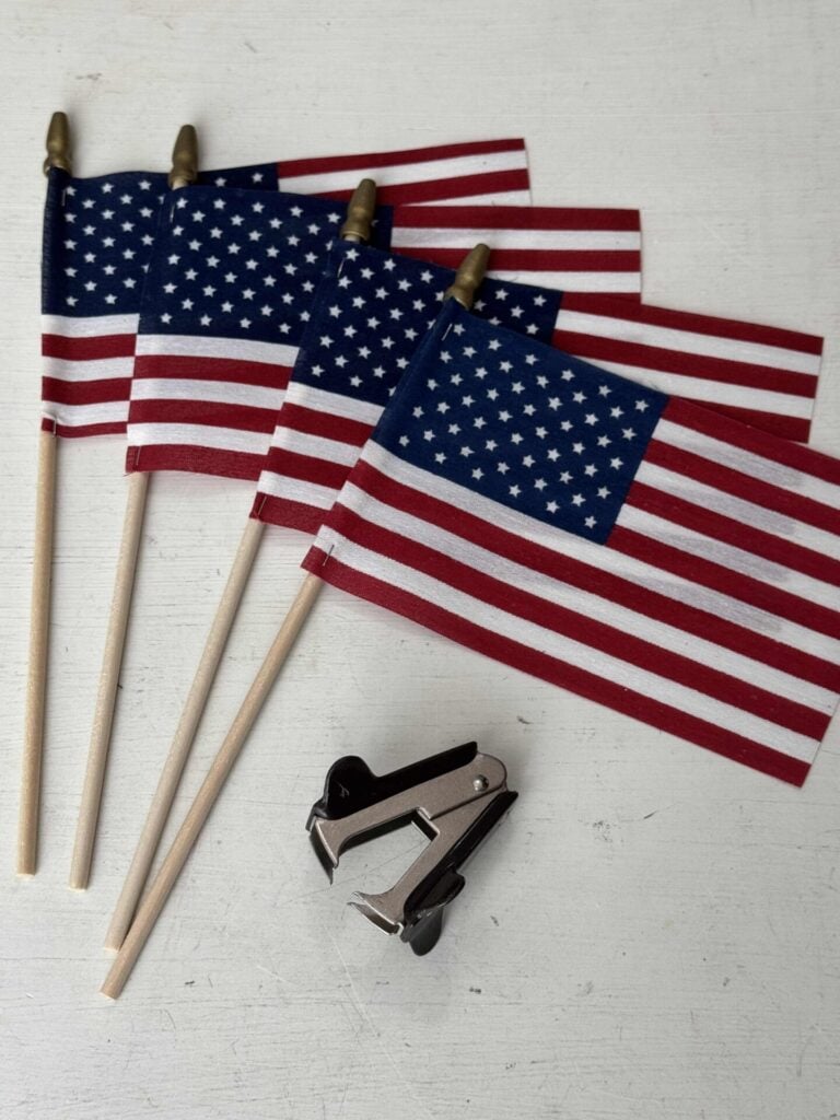 Four small American flags on wooden sticks are arranged on a white surface beside a metal staple remover, perfect supplies for crafting a DIY patriotic garland.