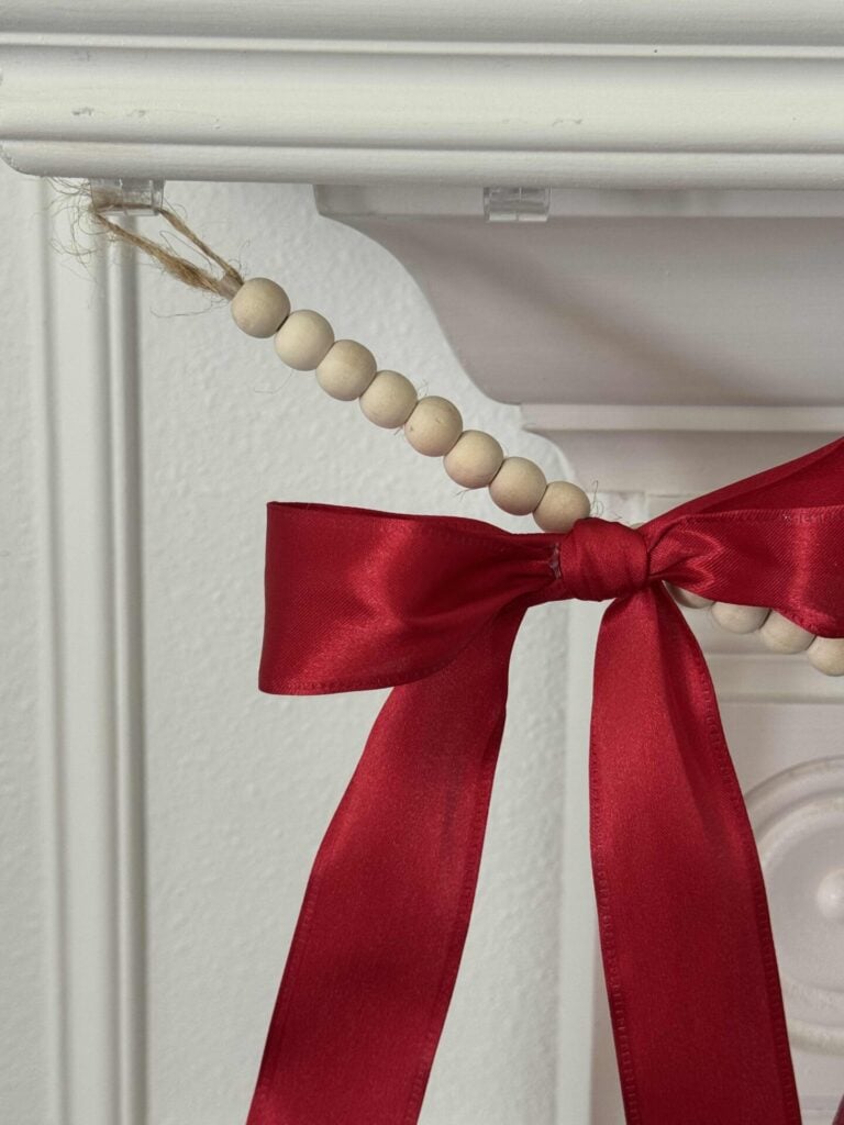 A close-up of a white fireplace mantle decorated with a DIY patriotic garland made from wooden beads and finished with a large red satin bow tied around it.