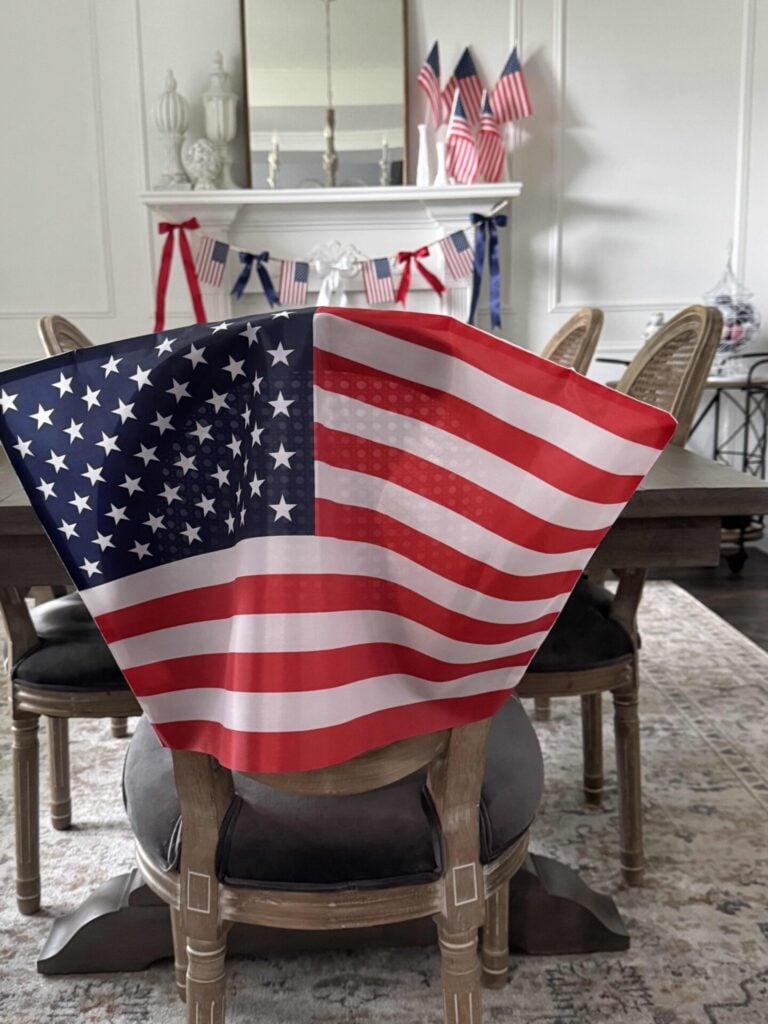 A wooden dining chair with an American flag draped over its back sits at a dining table. In the background is a mantel with a red, white, and blue ribbon garland and several small American flags.