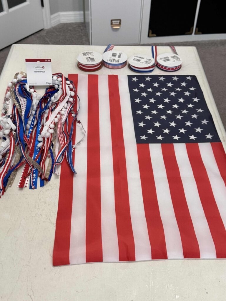 Red, white, and blue craft ribbons, an American flag, and spools of patriotic ribbon are arranged on a white table.