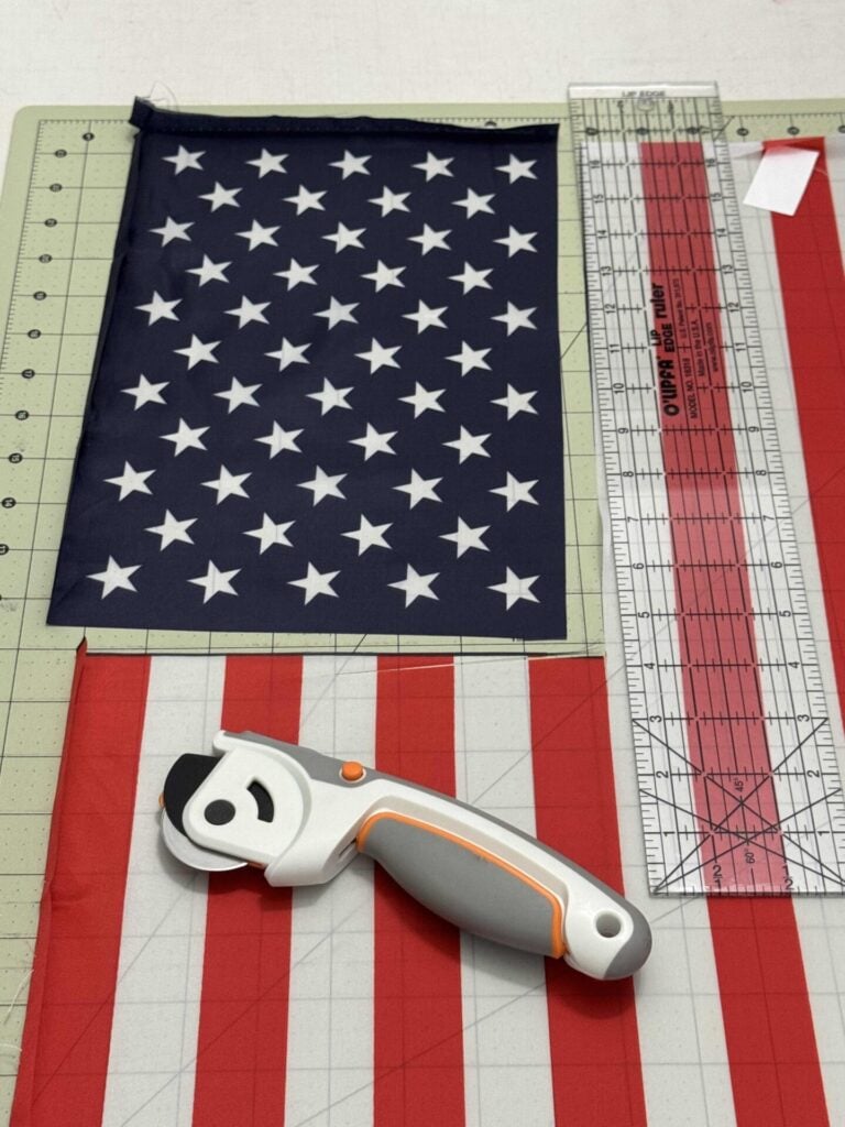 A rotary cutter, clear ruler, and cutting mat are laid out with red, white, and blue star-spangled fabric pieces.