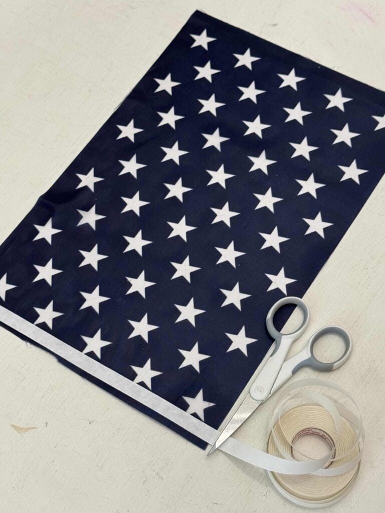 A piece of navy blue fabric with white stars, lies on a table beside a pair of gray scissors and a roll of double-sided tape.