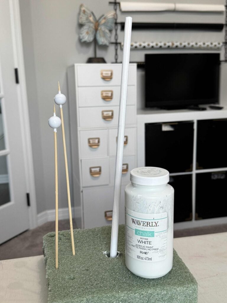 A bottle of Waverly white chalk paint sits on green foam, with a painted dowel and two beads drying upright. White storage cabinets and butterfly wall decor add charm to the background.