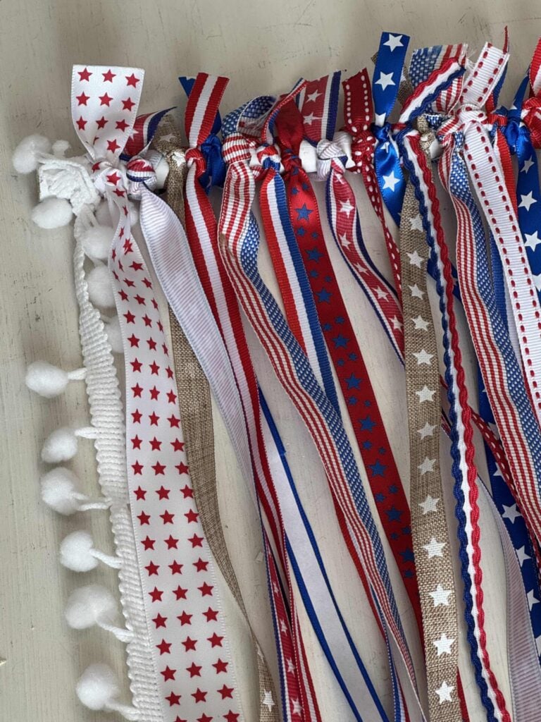 A close-up of ribbons tied on to a white dowel rod. On the left, a white trim with pom-poms is also visible.