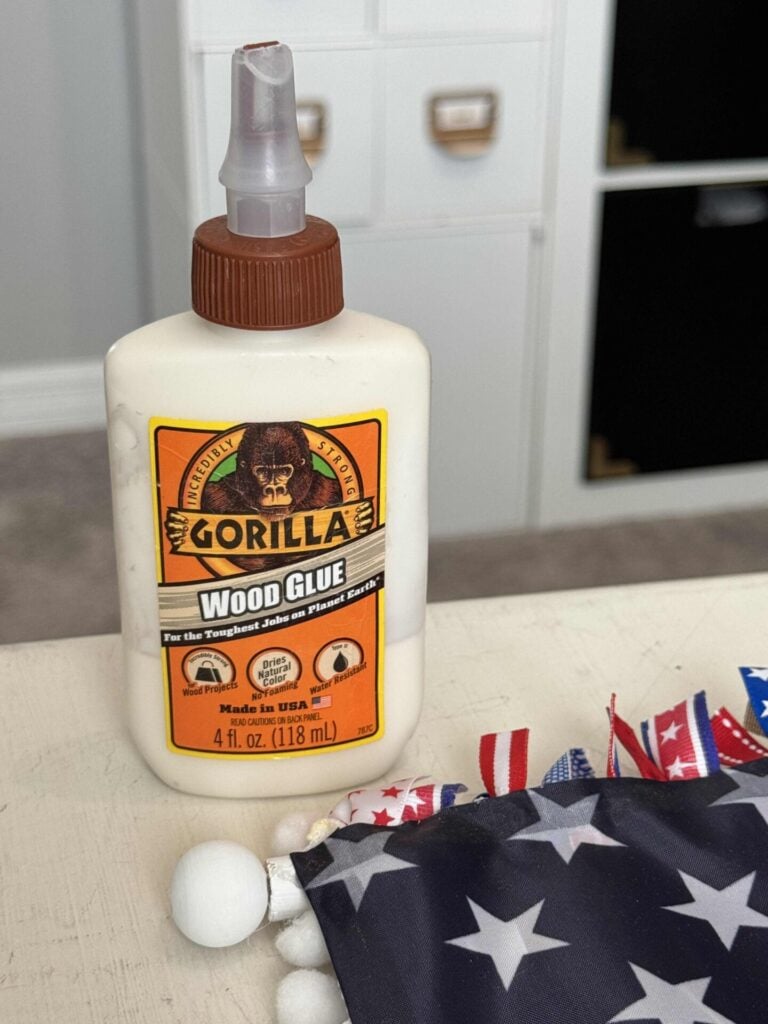 A bottle of Gorilla wood glue sits on a table next to a partially visible American flag-themed fabric with pom-pom trim, perfect for your DIY Patriotic Ribbon Flag project. Filing cabinets and shelves appear in the blurred background.