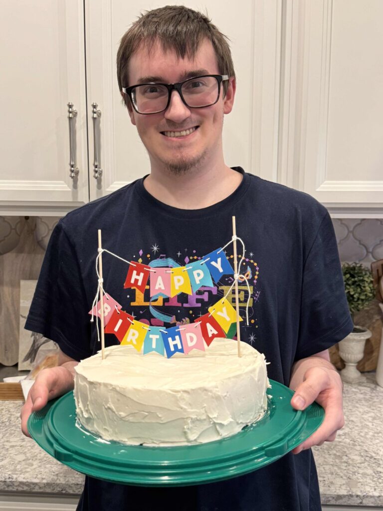 A person with glasses is smiling and holding a white frosted cake on a green plate. The cake has a colorful Happy Birthday banner on top, and they are standing in a kitchen.