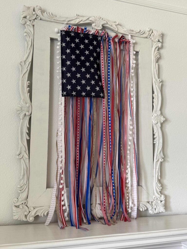 A decorative DIY Patriotic Ribbon Flag features an American flag section with stars and long red, white, and blue ribbons, displayed on a white ornate frame against a light-colored wall.