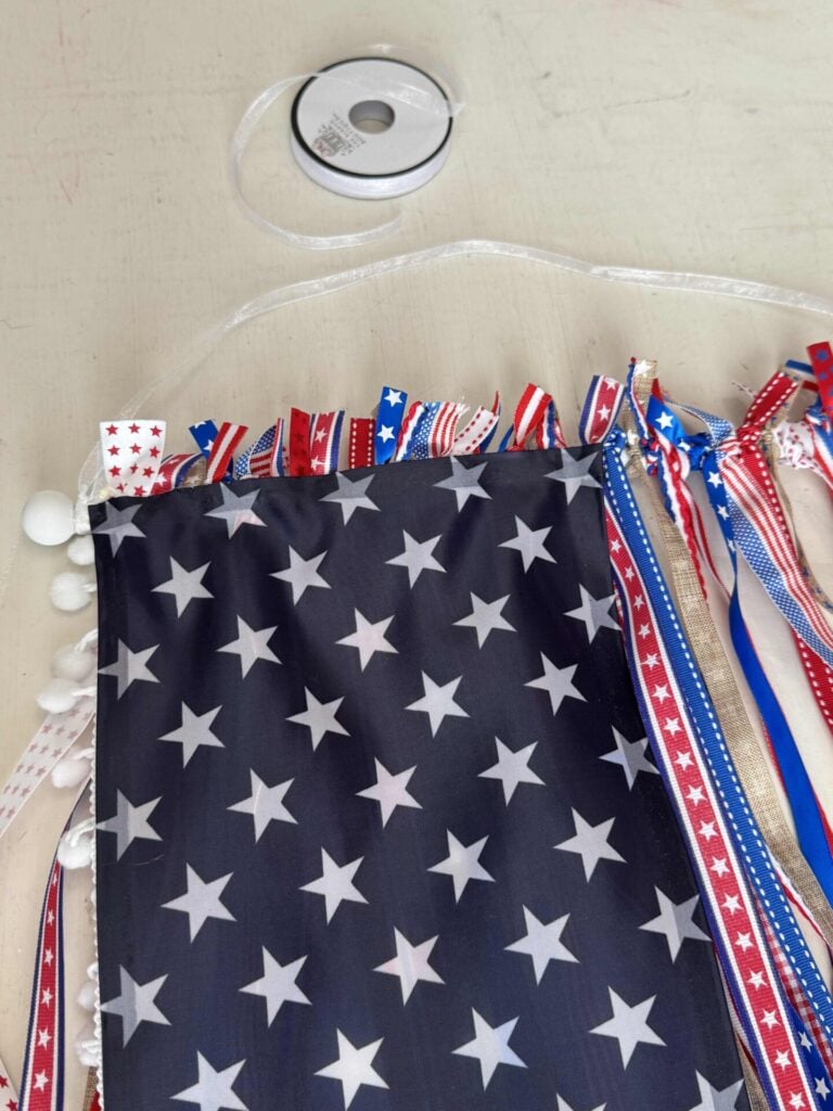 A close-up of a DIY Patriotic Ribbon Flag shows dark blue fabric with white stars, adorned with red, white, and blue ribbon strips and pom-poms, plus a spool of ribbon on a light-colored surface.