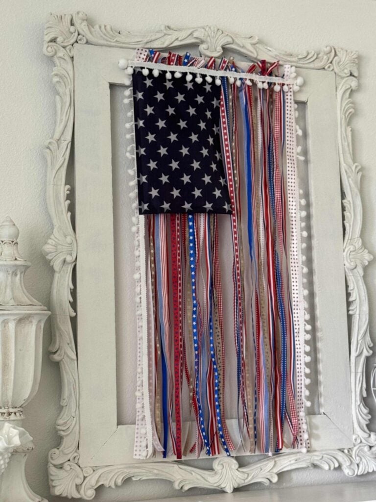 A framed DIY Patriotic Ribbon Flag features a blue field with white stars and red, white, and blue ribbons hanging vertically as stripes, all set against a white ornate frame on a light-colored wall.