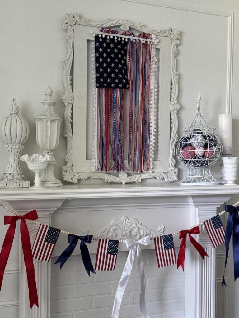 A white mantel decorated with American flags, red, white, and blue ribbons, and patriotic garlands. Milk glass vases and decorative finials sit on the mantel, with a framed DIY Patriotic Ribbon Flag display above.
