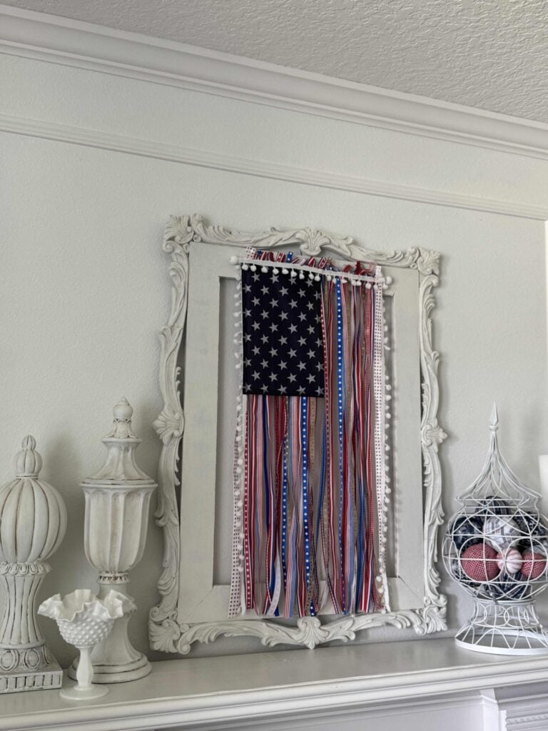 A decorative white-framed mirror displays a DIY Patriotic Ribbon Flag with hanging red, white, and blue ribbons. On the mantel are white statue finials, a milk glass dish and a wire basket with fabric orbs.